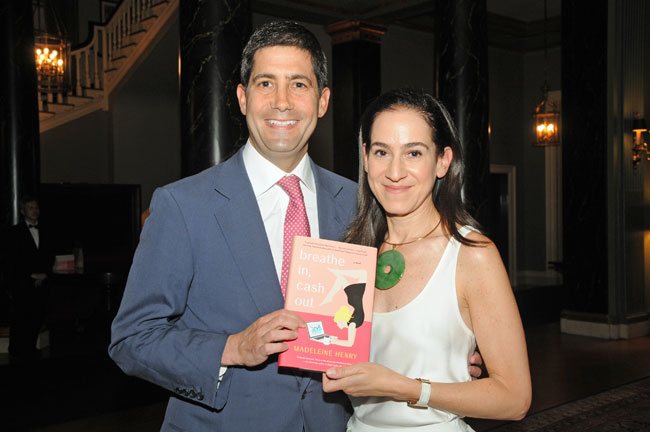 Kevin Warsh and Jane Lauder posing together indoors at an evening event, smiling and holding a copy of Breathe In, Cash Out — The Coinomist.