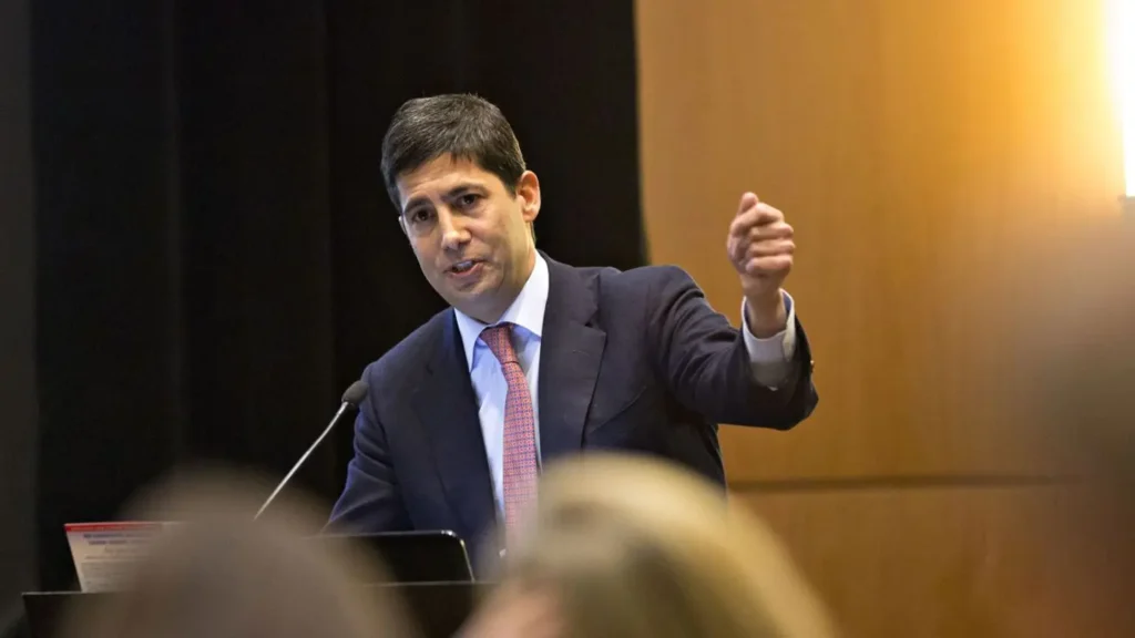 Kevin Warsh speaking from a podium with a raised hand during a public event — The Coinomist.