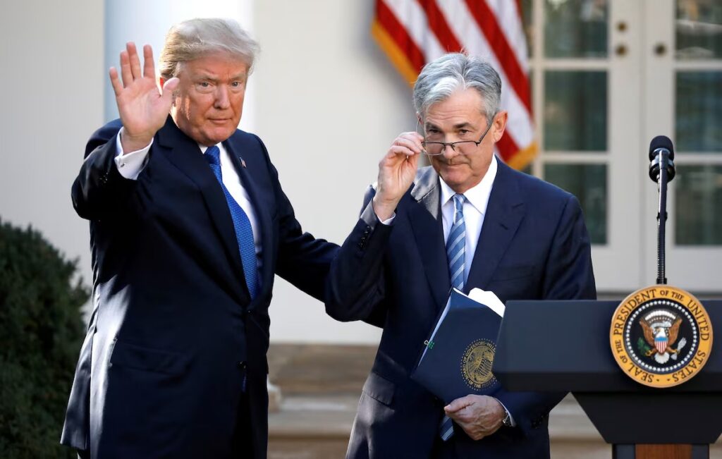 Donald Trump and Jerome Powell standing together at the White House podium, Powell holding a blue folder with the Fed seal — The Coinomist.