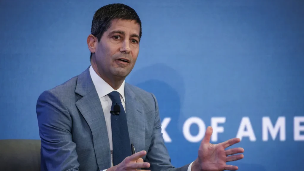 Kevin Warsh gesturing while speaking onstage at a financial summit — The Coinomist.