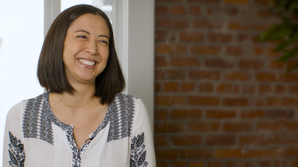 Monica Long smiling during an interview, wearing a patterned white blouse with a brick wall background — The Coinomist