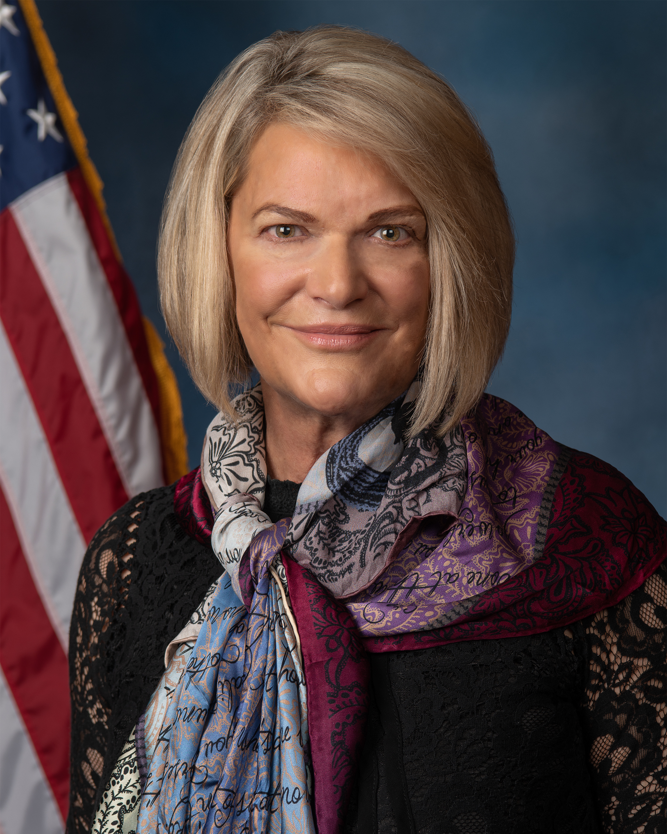 A photo of U.S. Senator Cynthia Lummis with a dark blue background and the U.S. flag - The Coinomist