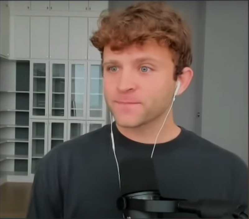 Jack Mallers speaking into a microphone with earbuds in, seated in front of glass cabinets during a remote video interview — The Coinomist