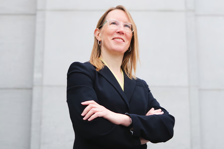 Hester Peirce standing with arms crossed, wearing glasses and a dark blazer, looking upward in front of a stone wall — The Coinomist