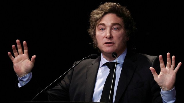 Javier Milei standing with raised hands at a podium, speaking before an audience, photographed during official statements. — The Coinomist