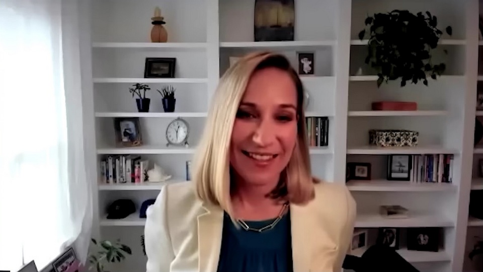Kara Calvert smiling during a virtual interview, seated in front of white bookshelves — The Coinomist