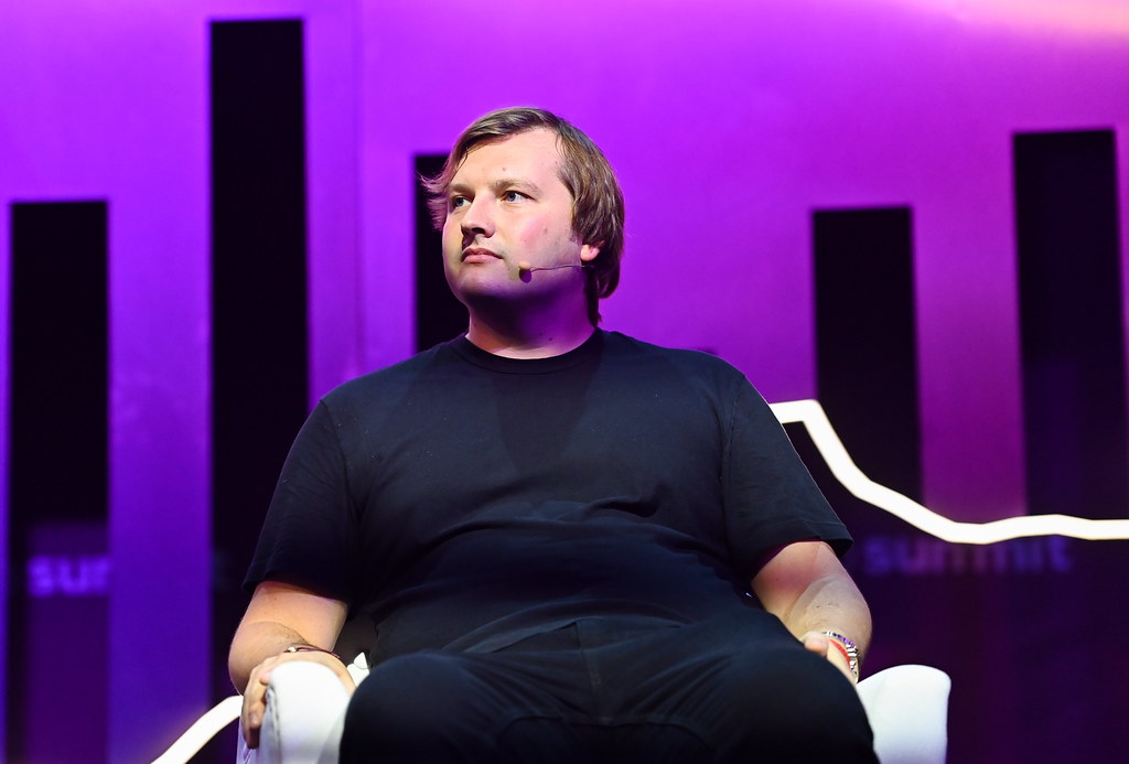 Stani Kulechov seated on stage under pink lighting, wearing a black t-shirt and headset microphone, gazing to the side during a conference — The Coinomist