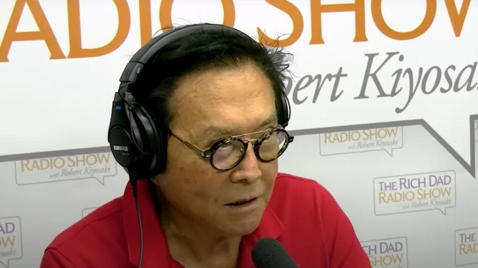 Robert Kiyosaki speaking into a microphone during a recording of his Rich Dad Radio Show, wearing glasses and a red shirt — The Coinomist