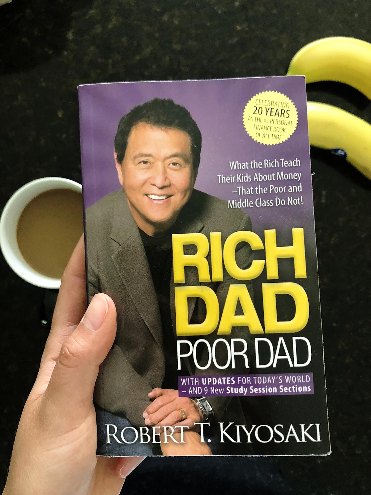 A reader holding a copy of Robert Kiyosaki’s book Rich Dad Poor Dad, with Kiyosaki smiling on the purple cover next to a cup of coffee — The Coinomist
